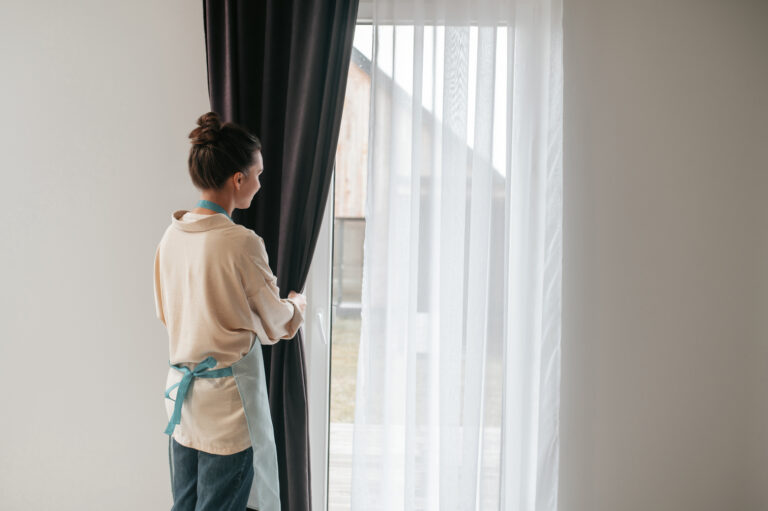 Daily routine. Young woman standing near the widnow and fixing the curtains